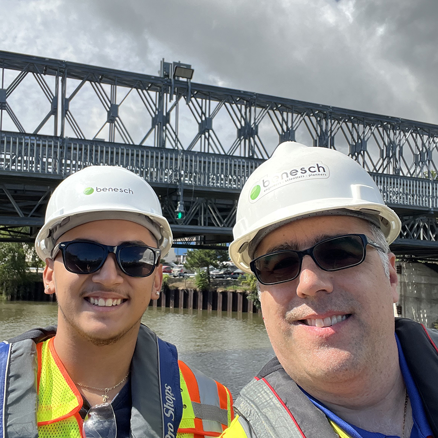 A photo of Juan an intern and Mark an engineer in the field smiling in front of the bridge and wearing construction helmets