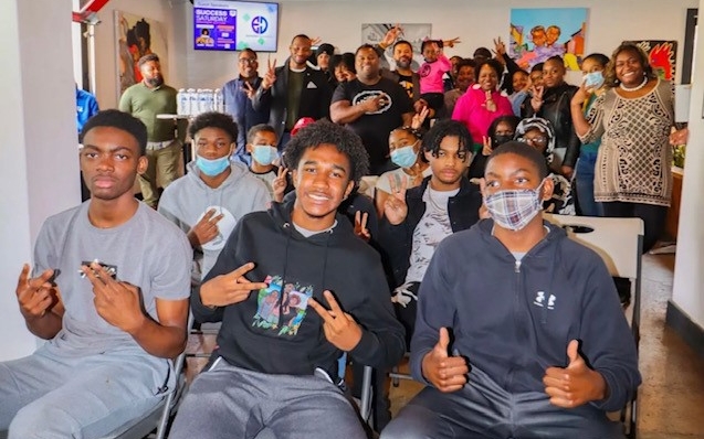 A photo of students in a classroom seated smiling towards the camera with thumbs up