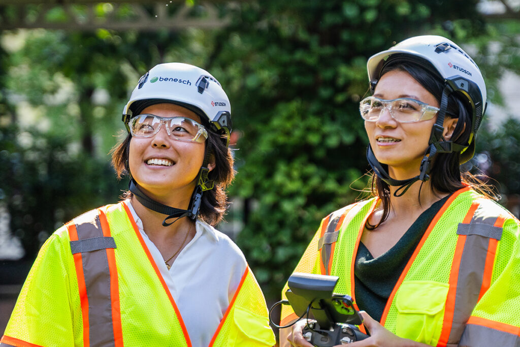 two Benesch engineers wear safety helmets and goggles as they fly a drone, which is off screen